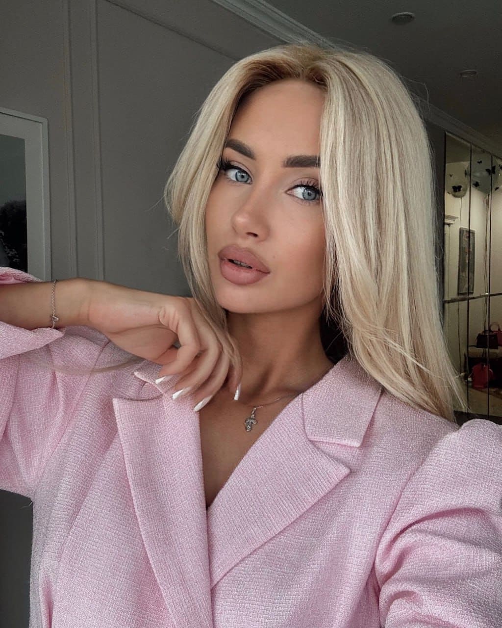 Vivienne stands indoors in a light pink textured blazer with a deep V-neck, raising her right hand near her collarbone.