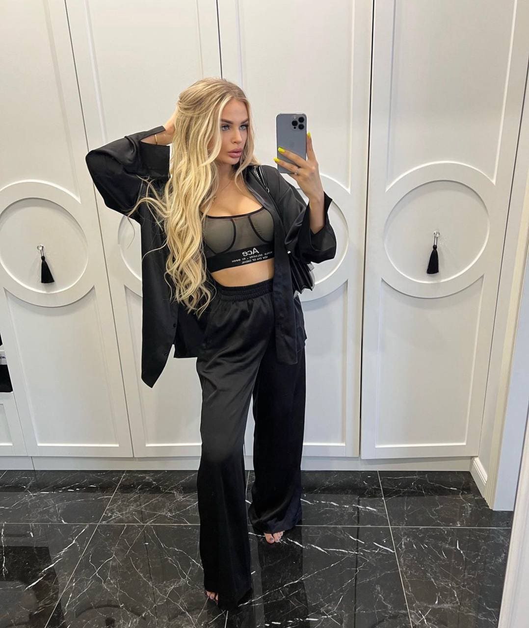 A mirror selfie shows a woman in a black satin jacket and wide-leg pants, holding a phone, against white paneled walls.