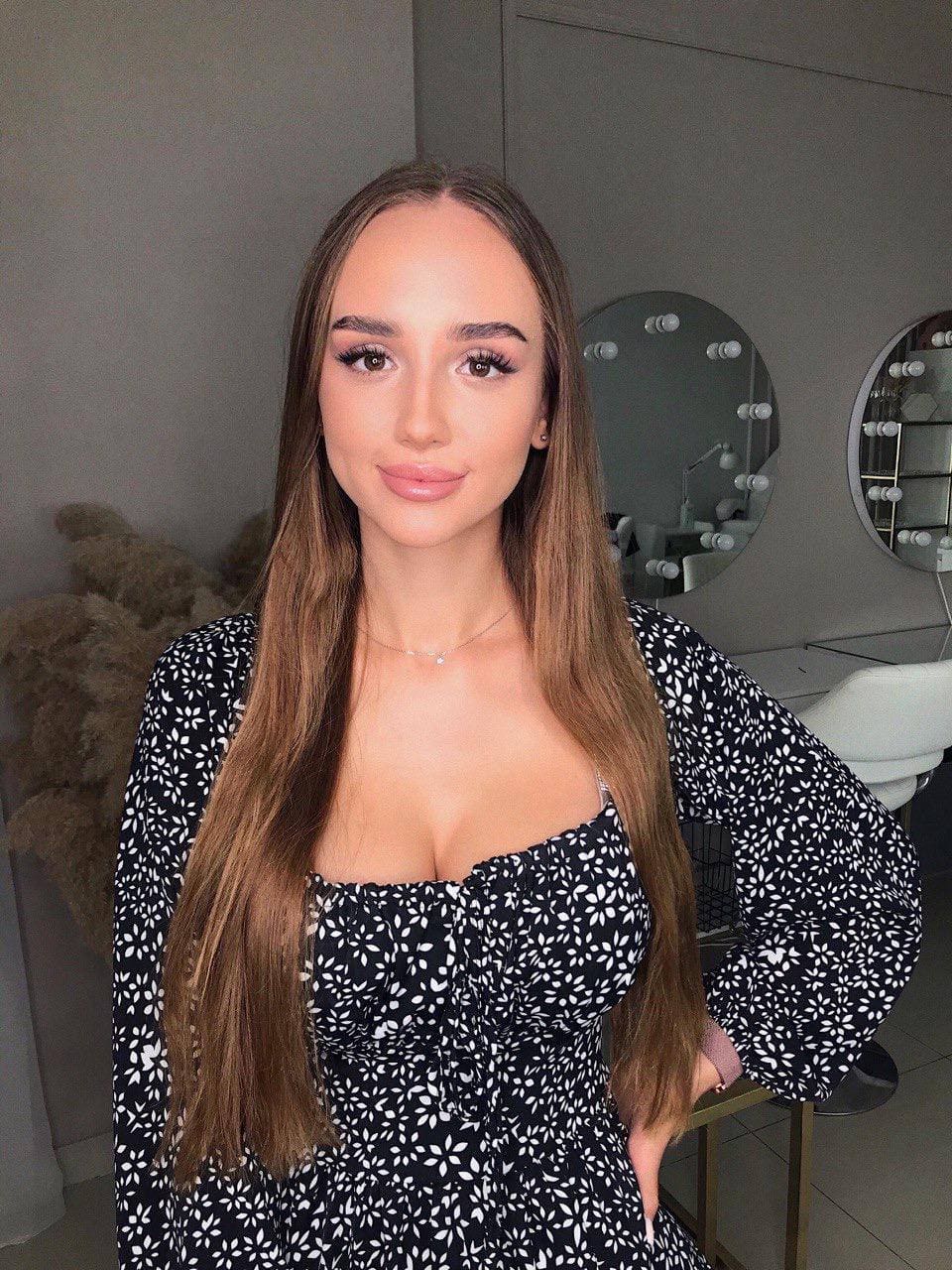 Elichka stands facing the camera in a black and white floral dress with a low neckline, in a room with round mirrors and a white chair.