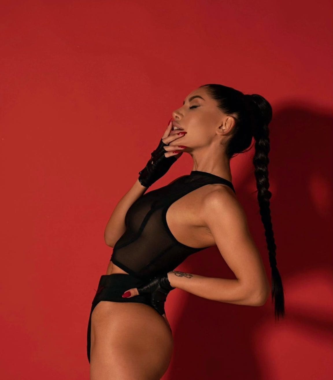In profile against a red wall, a woman with a long braid wears a sheer black crop top, high-cut bottoms, and fingerless gloves.