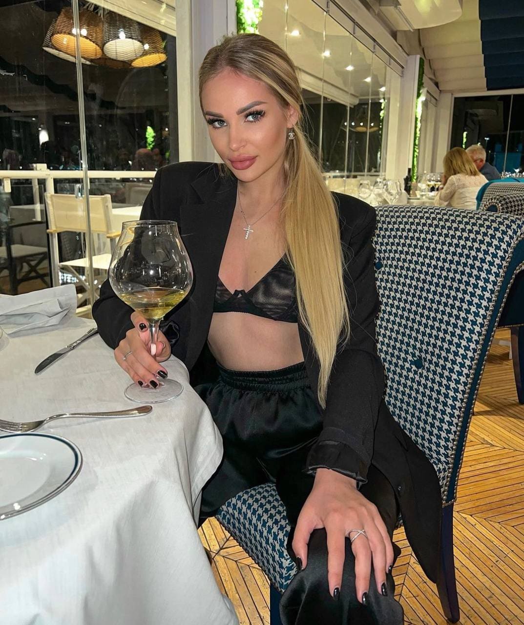 Lena sits at a restaurant table in a black blazer, bra, and satin pants, holding a glass of white wine.