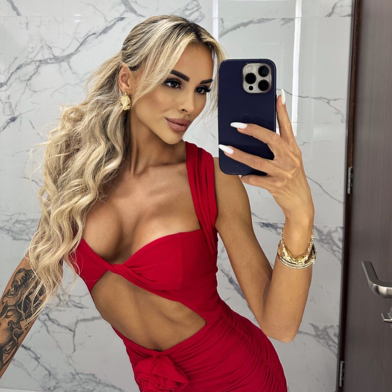 Tanya takes a mirror selfie in a red one-shoulder dress with side cutouts, standing before a marble-tiled wall.