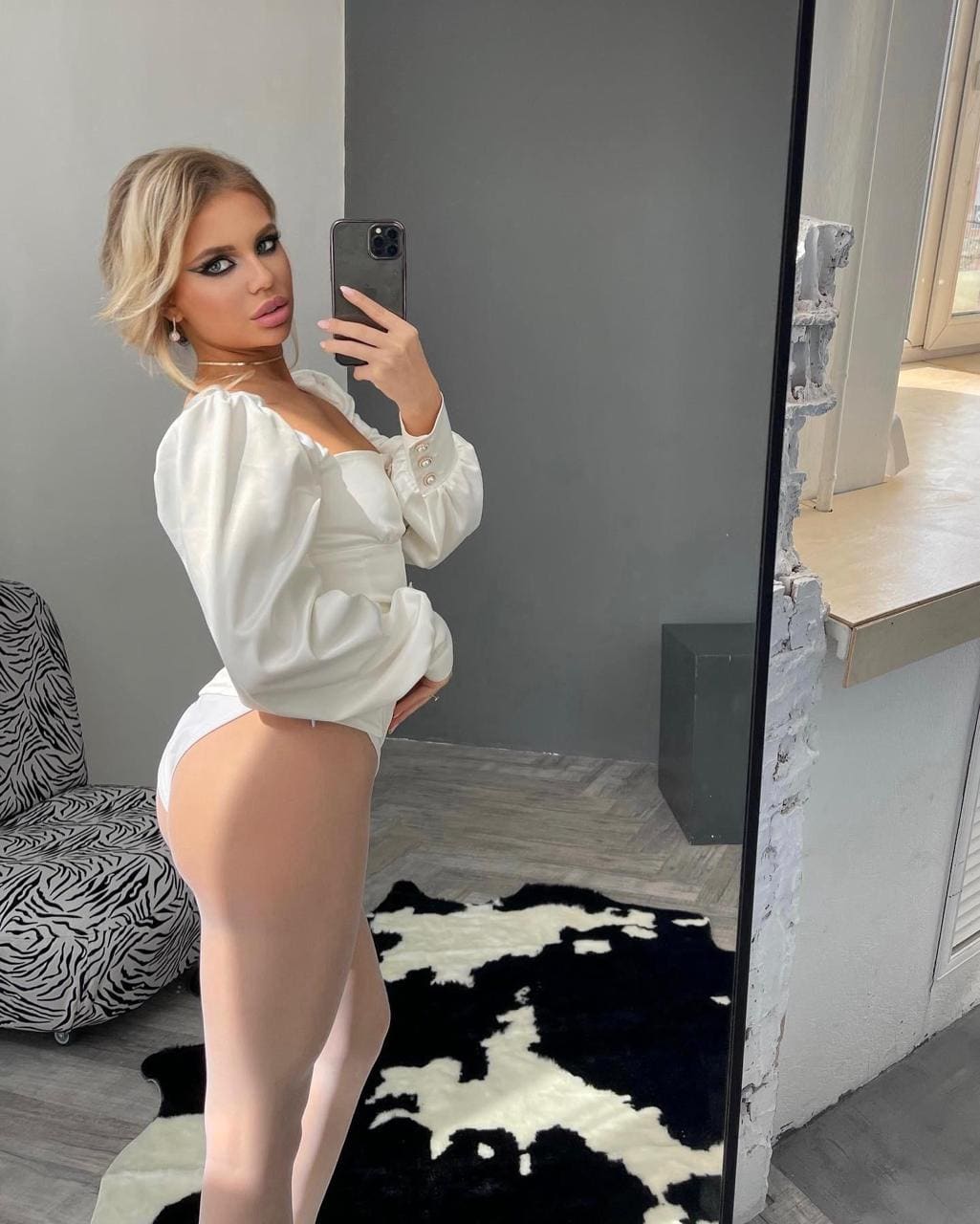 A woman with a short bob takes a mirror selfie in a white blouse and briefs, standing on a cowhide rug near a zebra-print chair.