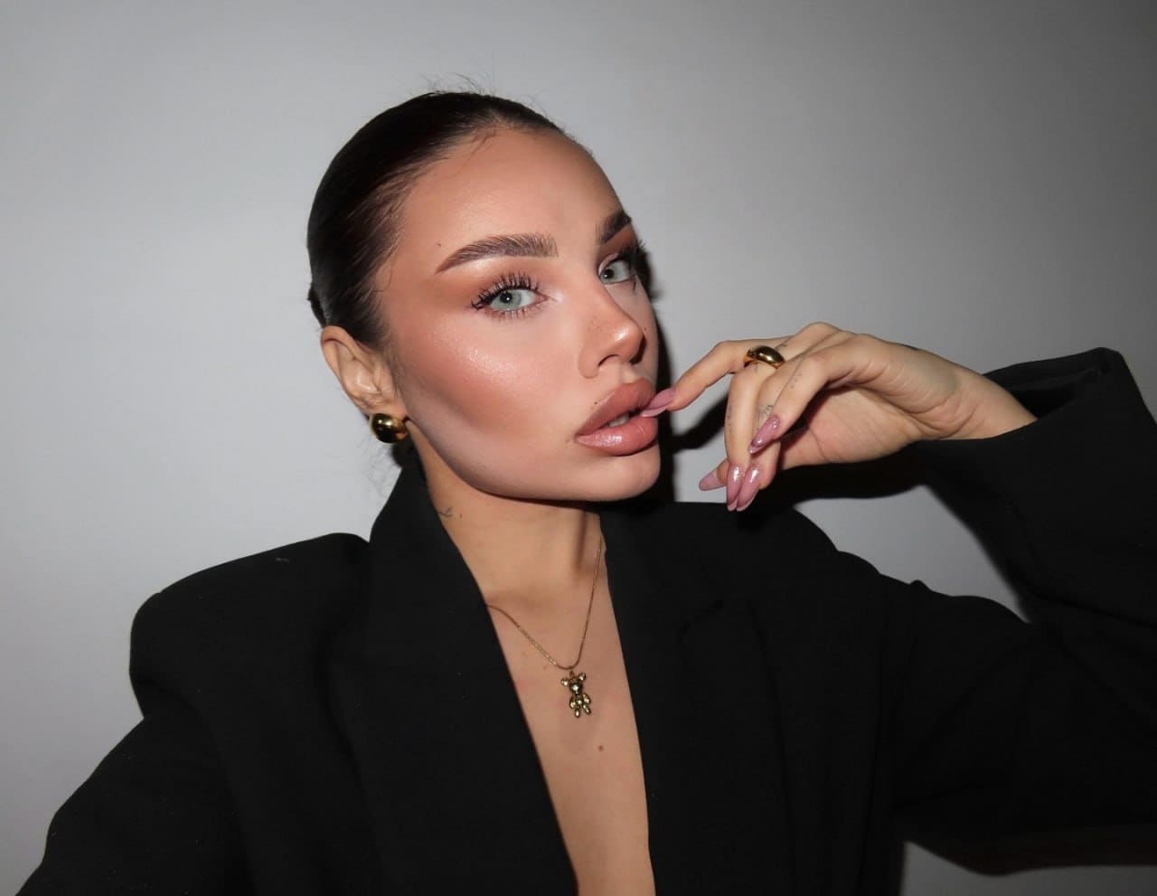 Alicia wears a black blazer with a deep V-neck and gold jewelry, posing with a hand near her lips against a gray background.