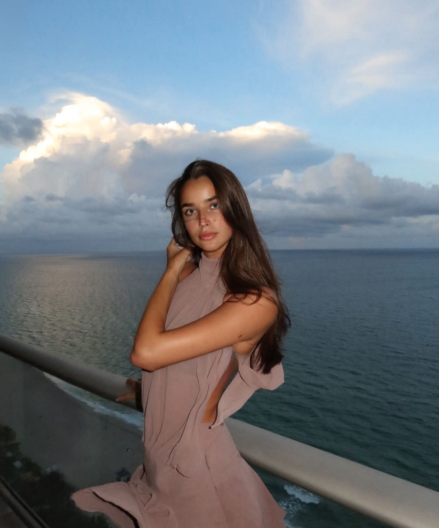 A woman in a dusty-pink cutout dress stands on a balcony overlooking the ocean, one hand raised to her hair.