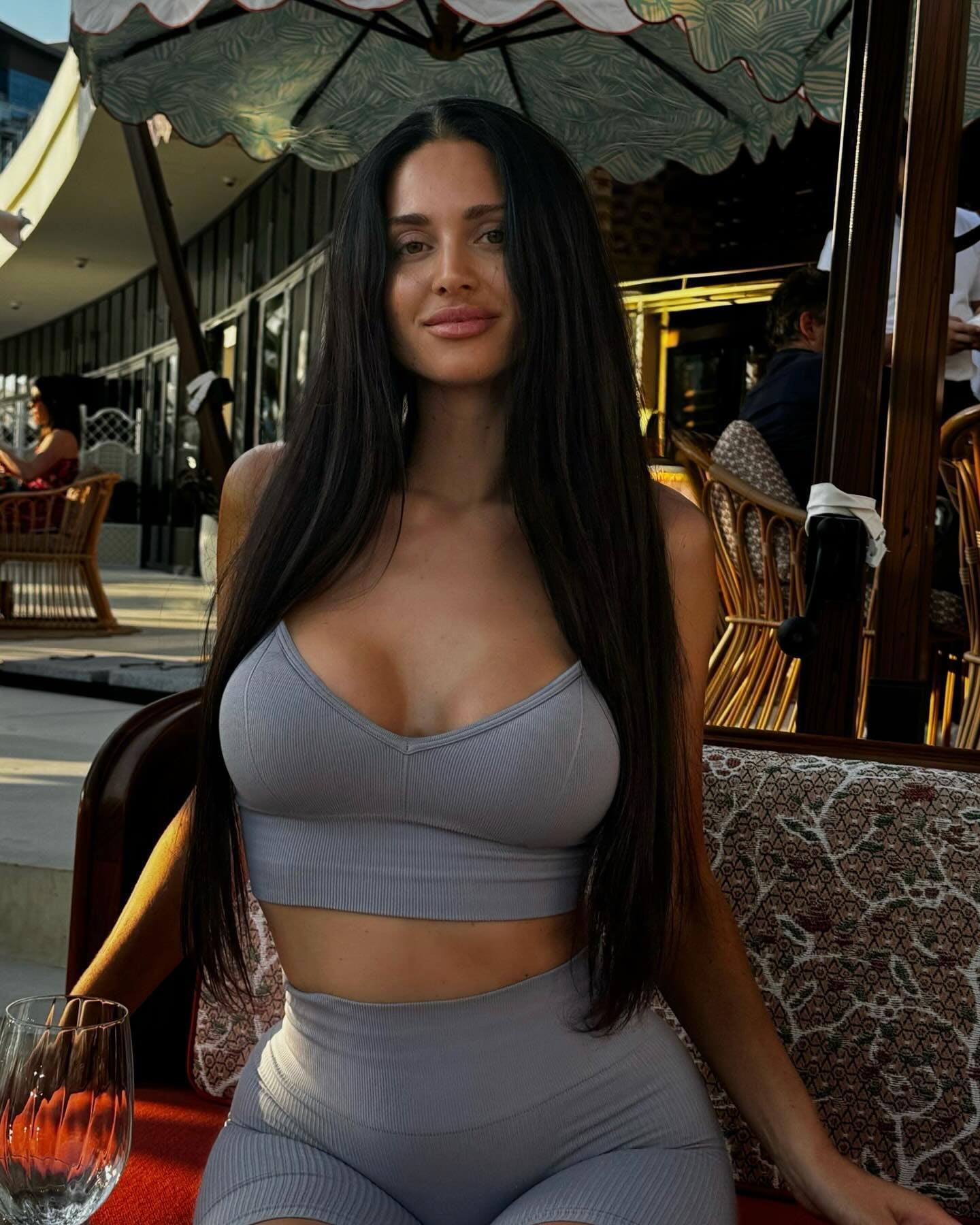 A woman in a grey ribbed crop top and leggings sits at an outdoor table under a patterned umbrella, with a glass of water nearby.