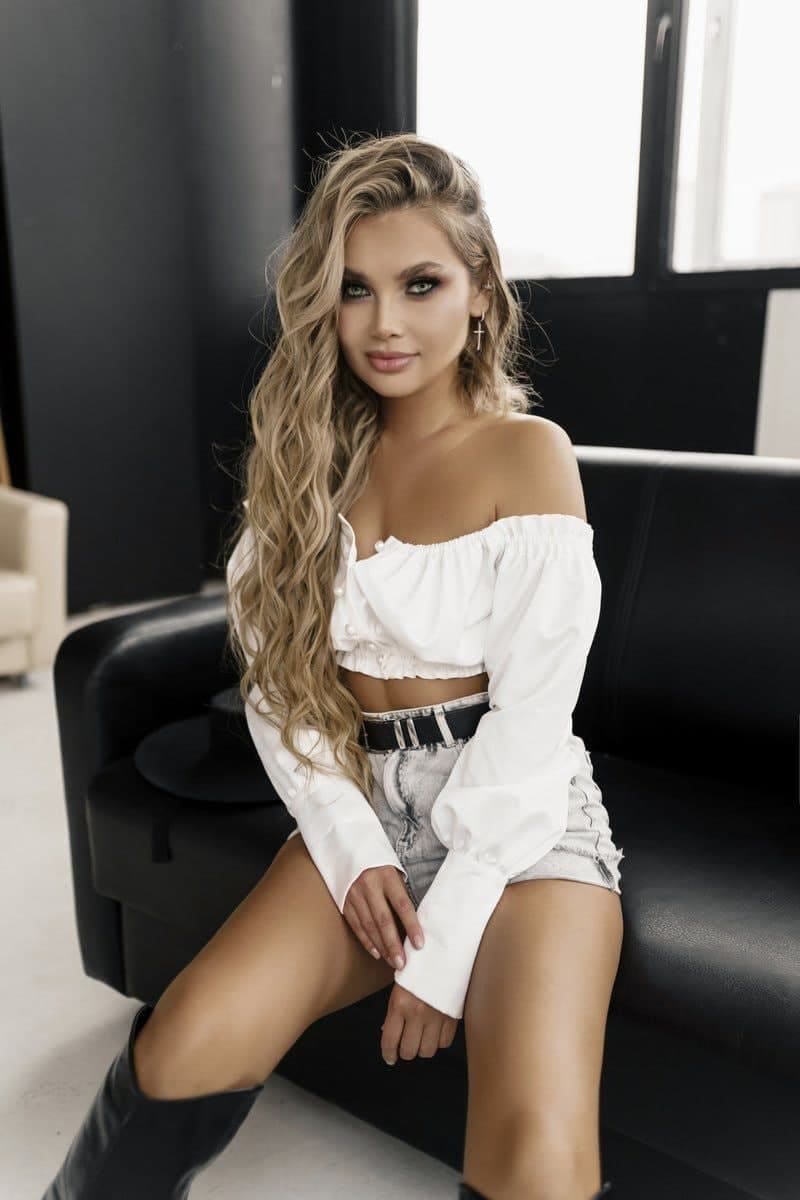 Ramina sits on a black leather couch in a white off-the-shoulder crop top, denim shorts, and black knee-high boots, with a window behind.