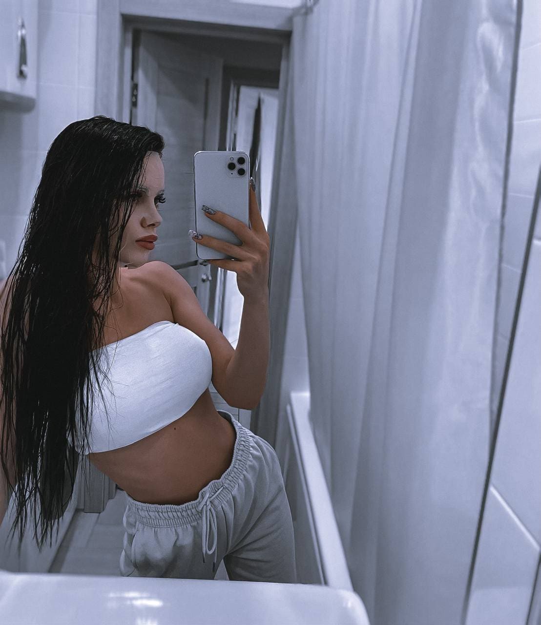A woman with wet hair takes a mirror selfie in a tiled bathroom, wearing a white tube top and grey sweatpants while holding a phone.