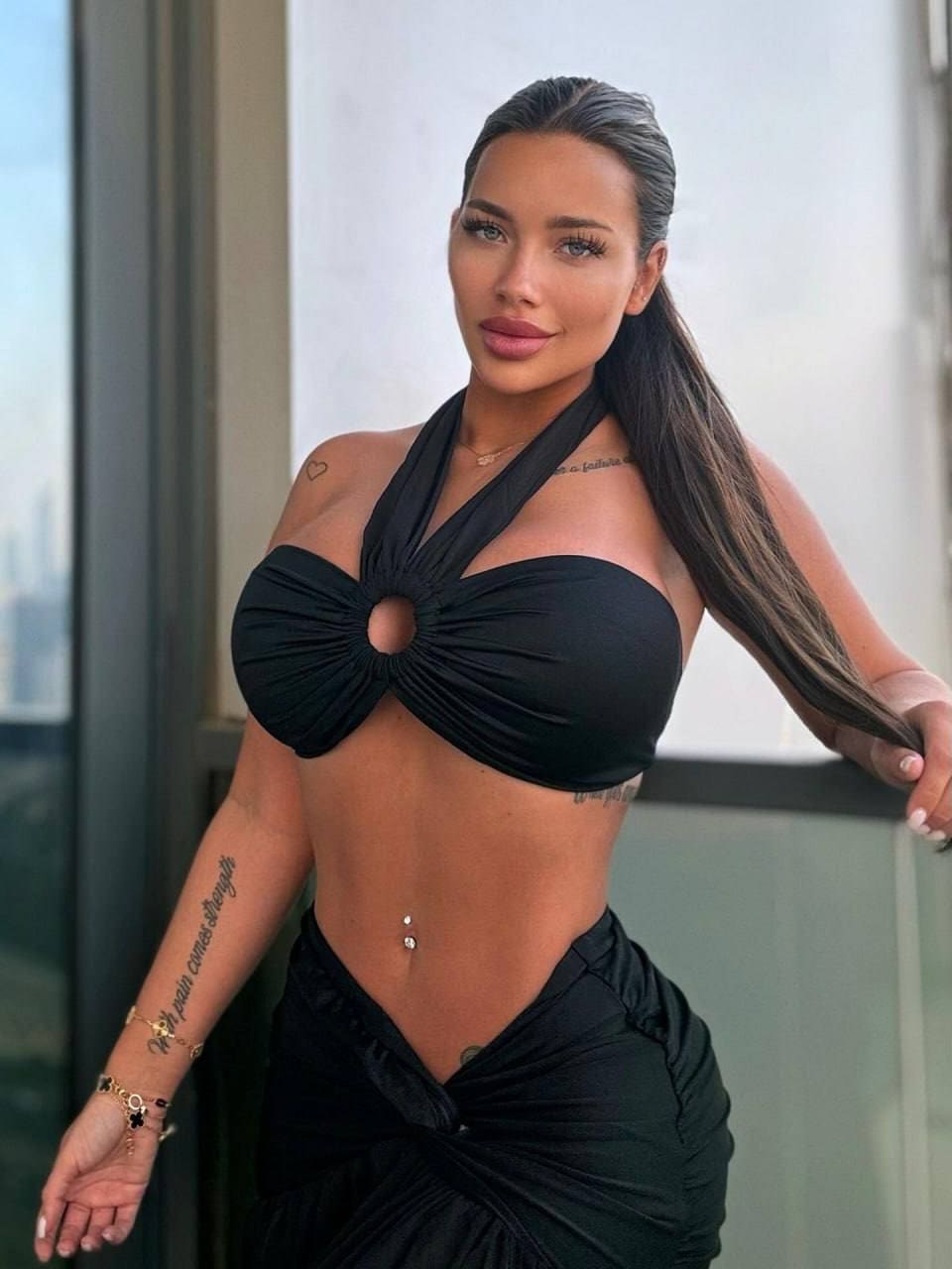 A woman with visible tattoos stands on a balcony in a black halter-neck cutout top and low-slung wrap skirt.