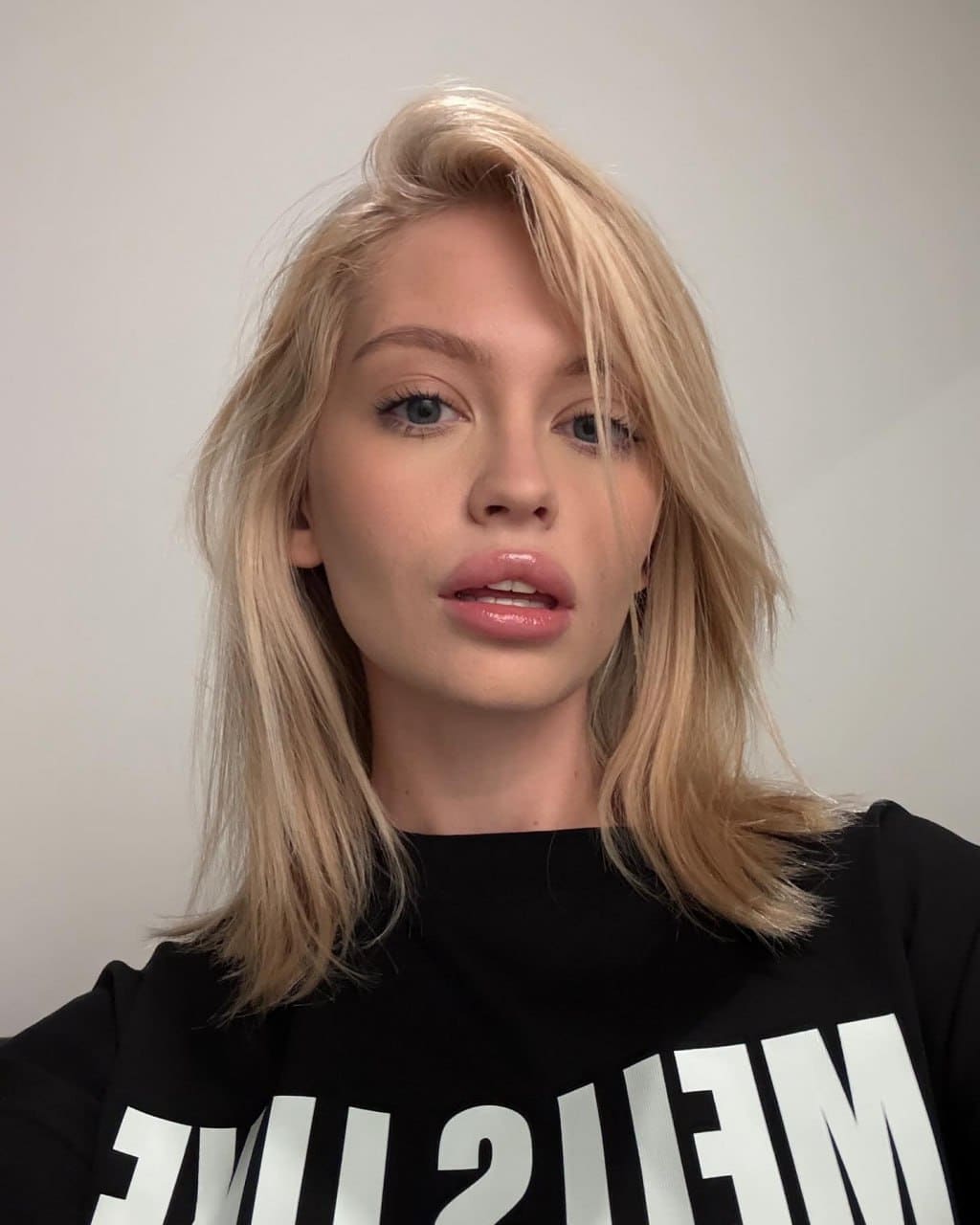 A woman with shoulder-length blonde hair wears a black sweatshirt with large white lettering, shown from the chest up against a light background.