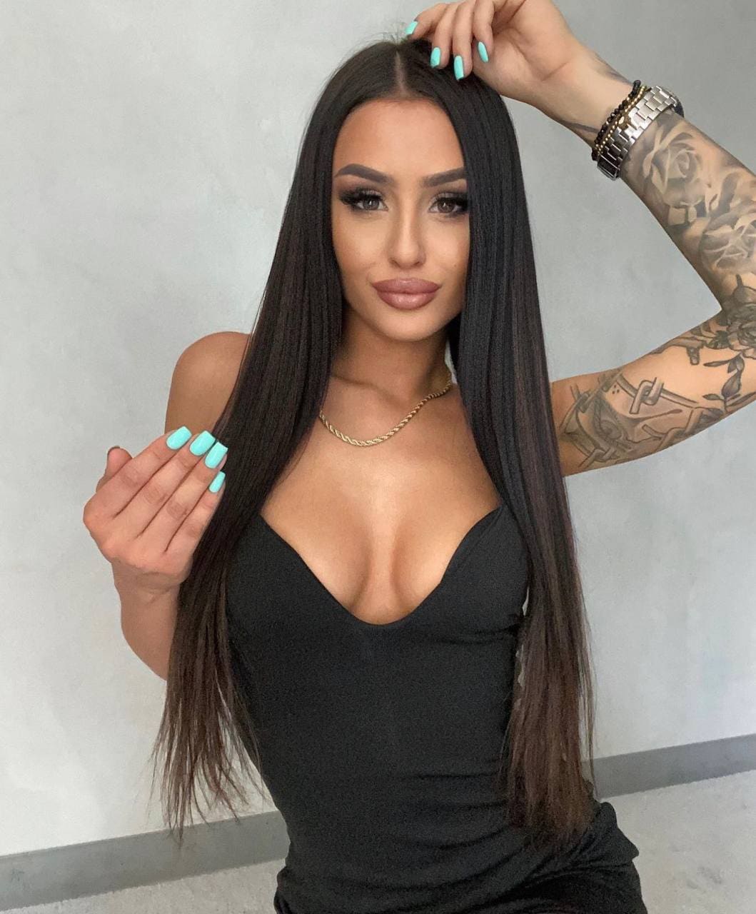 Amira with long dark hair and a tattooed left arm wears a black sleeveless top, one hand raised to her hair, against a plain light wall.