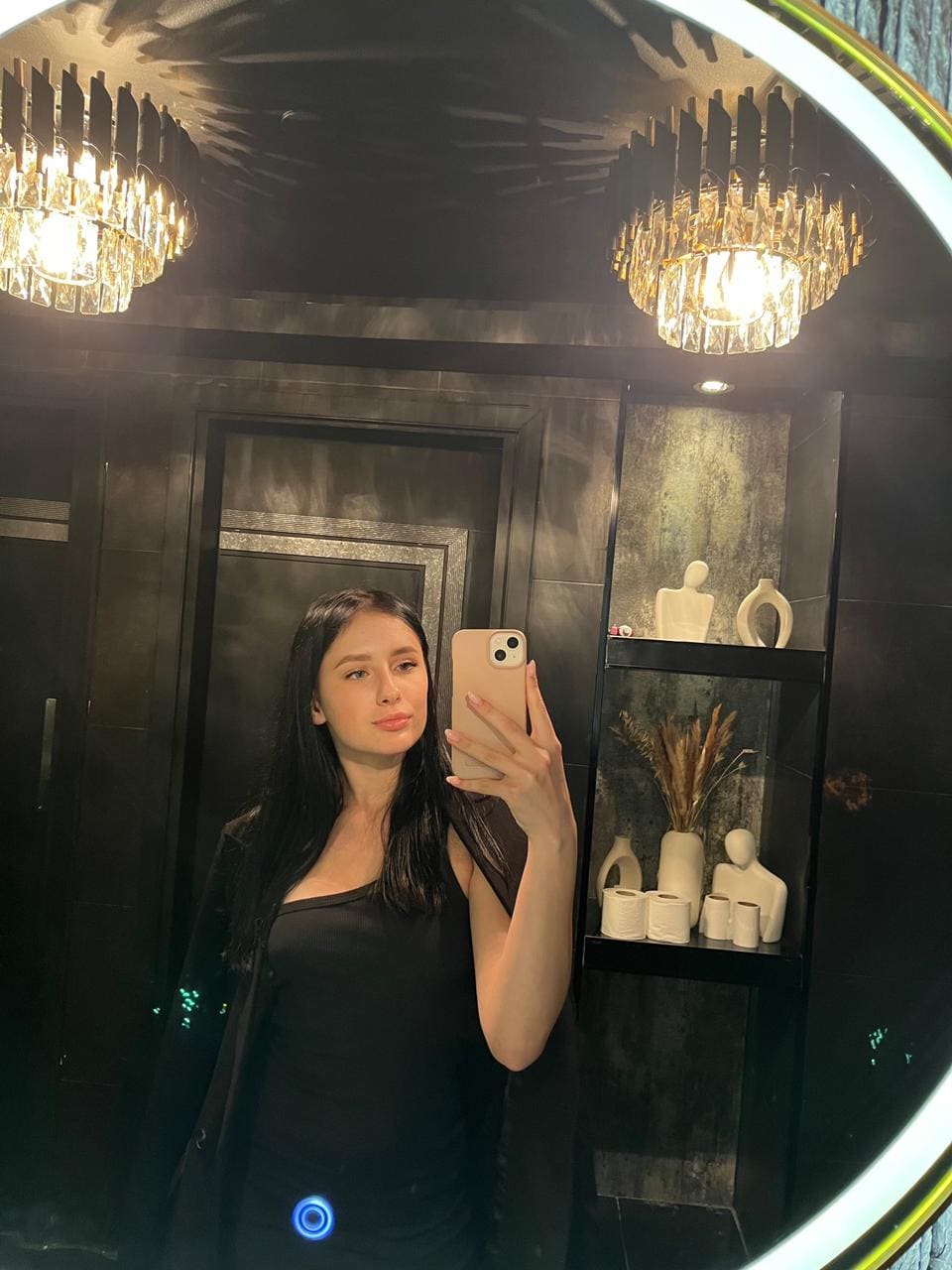Mirror selfie in a dark-tiled bathroom showing a woman in a black one-shoulder top and jacket, with a chandelier and shelf behind.