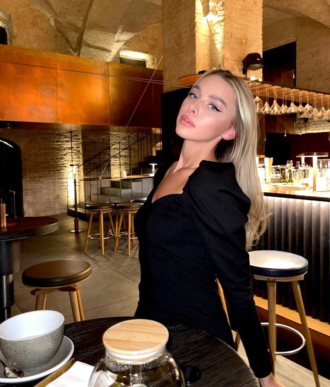 Zara sits at a dark wooden table in a restaurant, wearing a black long-sleeved top with a low neckline, with a bar and stools in the background.