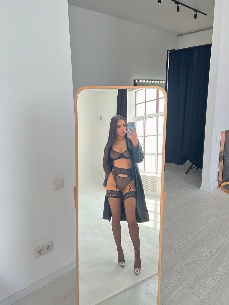 A woman in a black lace lingerie set and open dark coat takes a mirror selfie in a room with white walls and a window.