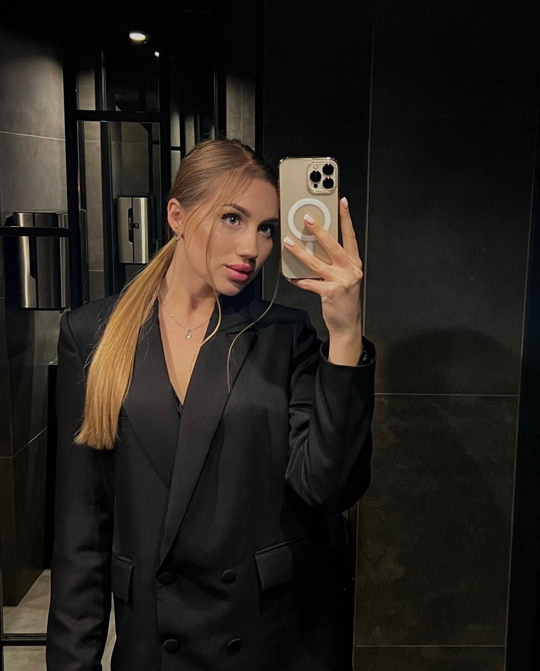 Woman with a side ponytail in a black double-breasted blazer, captured in a mirror selfie in a dark tiled room.