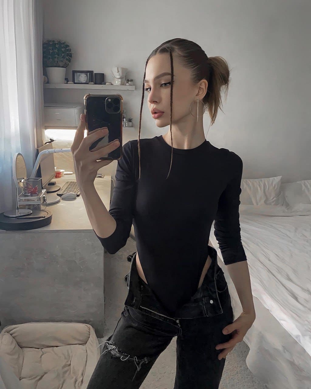 A mirror selfie in a bedroom shows a woman with a high ponytail in a black cutout top and distressed jeans, standing beside a bed and computer desk.