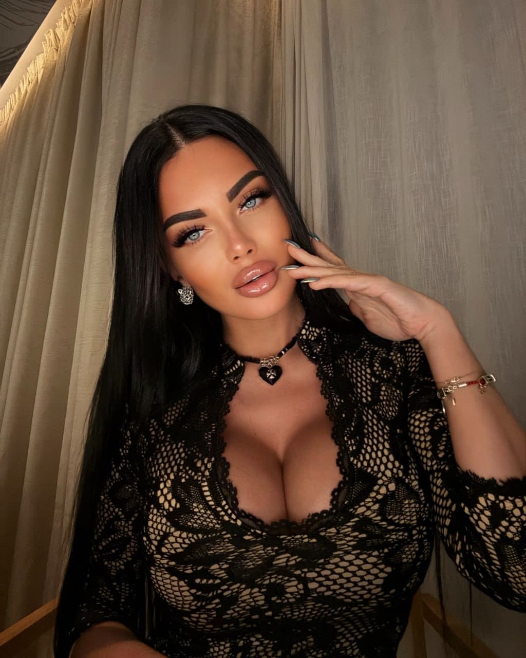 A woman in a black lace top and choker poses with her hand raised to her cheek in front of a beige curtain.