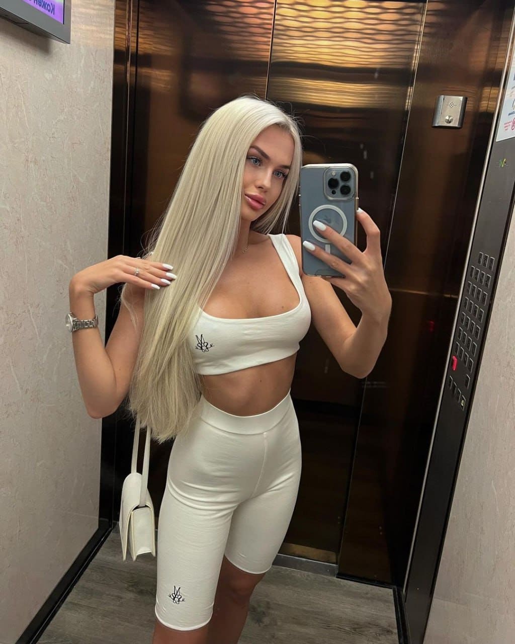 A platinum-blonde woman in a white crop top and shorts takes an elevator selfie holding a small bag.