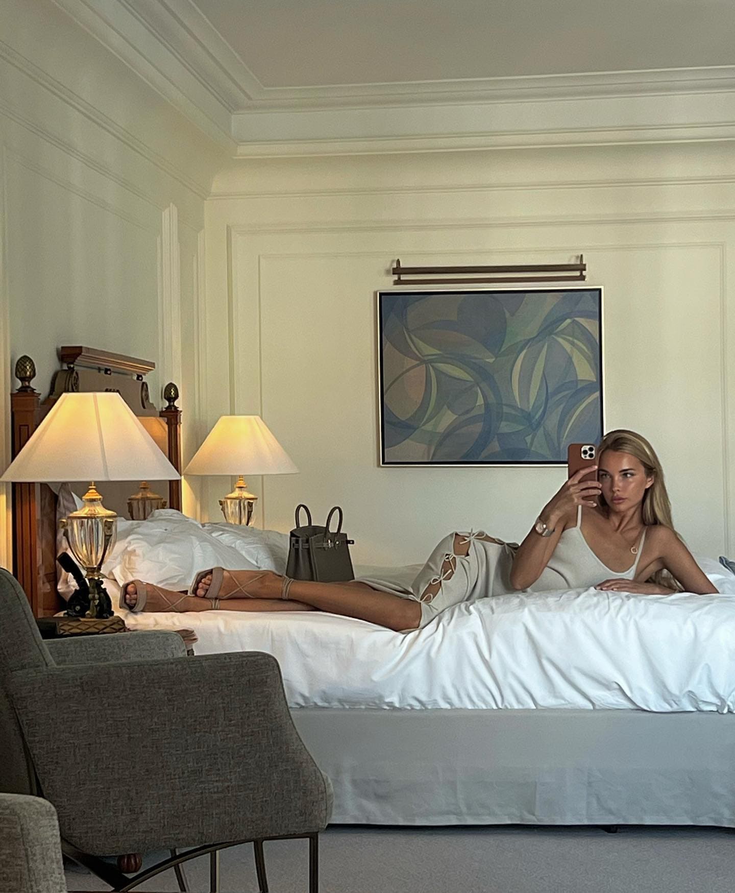 A blonde woman in a white sleeveless dress lies on a white bed, taking a mirror selfie with a phone, near lamps and abstract art.
