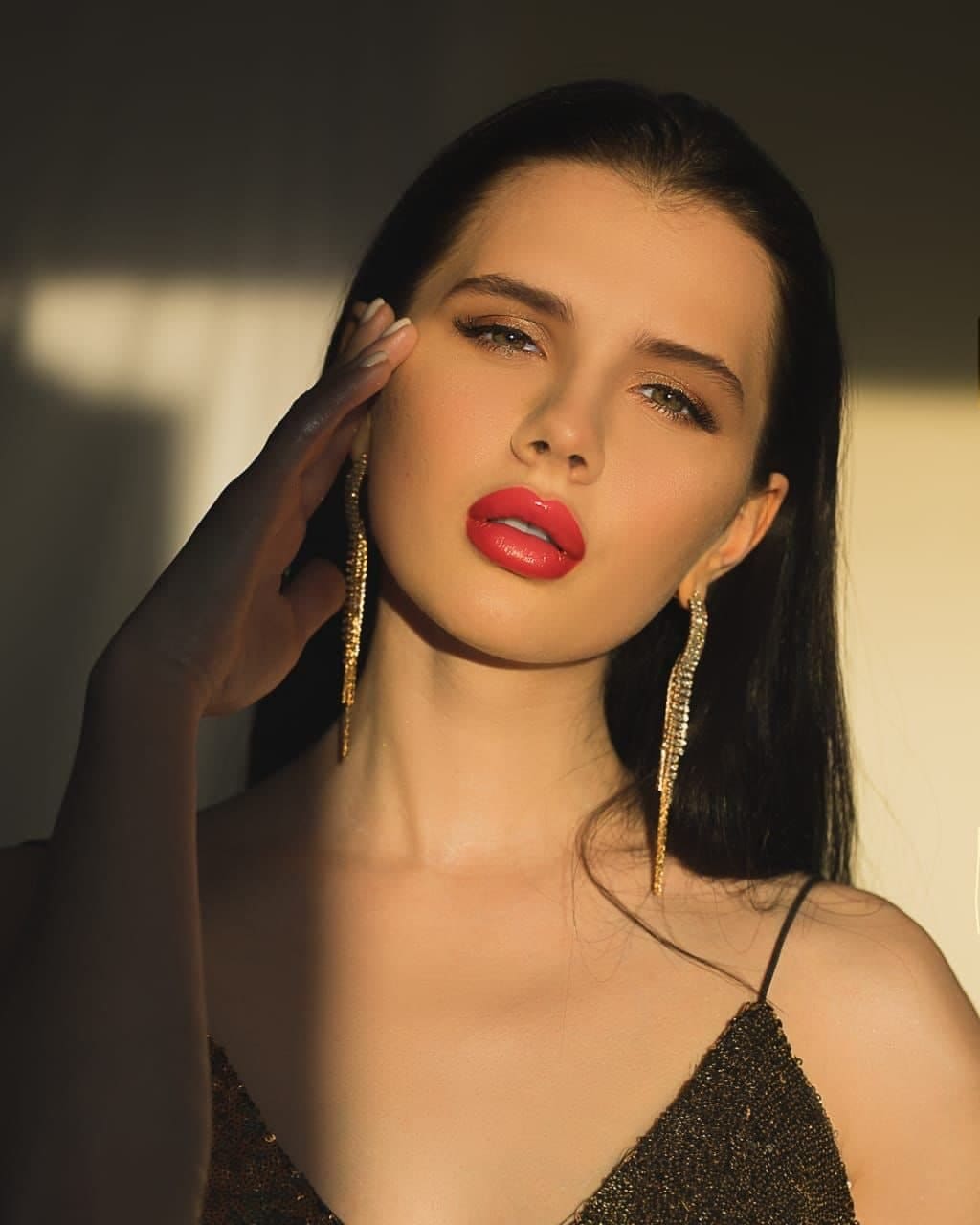 Daisy with long dark hair and red lipstick poses with a hand to her temple in a black sequined top and gold tassel earrings against a neutral background.