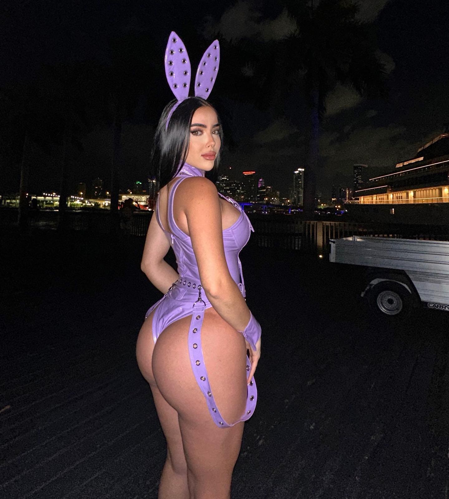 A woman stands in profile at night wearing a purple studded bunny-ear headband, a matching halter bodysuit with garter straps, and gloves, with a city skyline behind.
