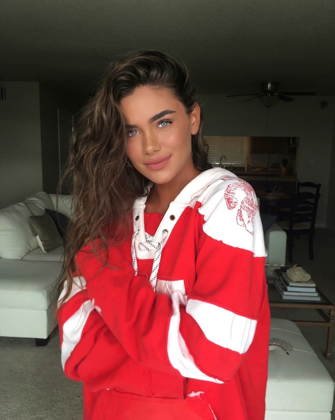 A young woman in a red and white hooded sweatshirt stands with arms crossed in a living room with a white sofa.