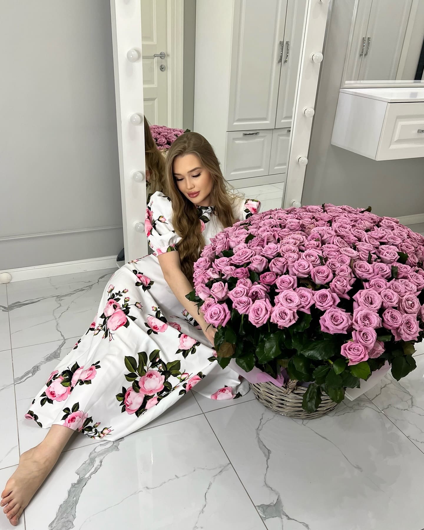 Woman in a white dress with pink roses sits barefoot on marble, leaning against a basket of flowers near a mirror.