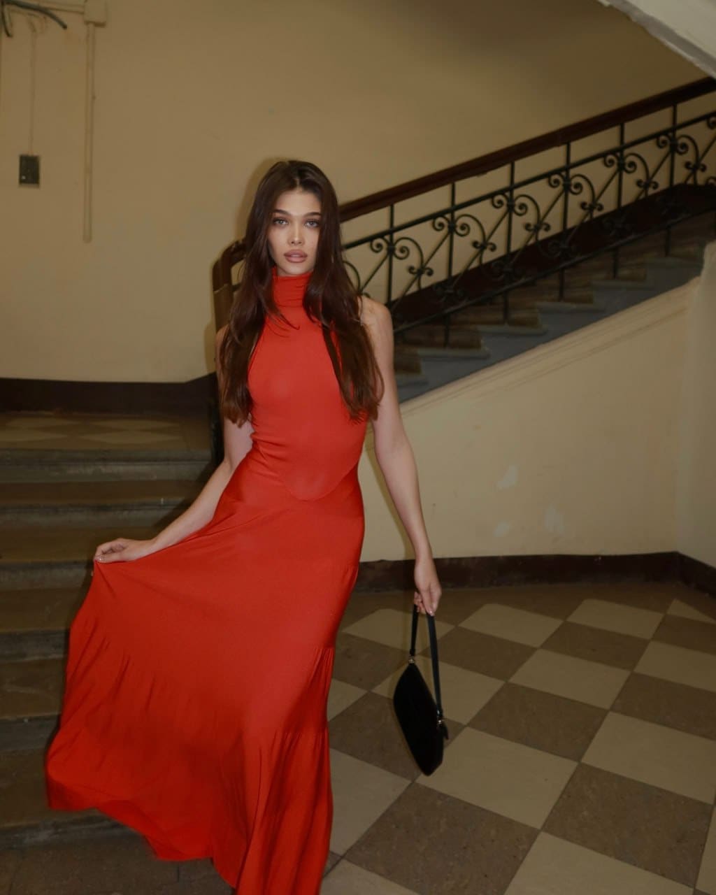 A woman in a red floor-length dress stands on a checkered floor near a staircase, holding a small black handbag and lifting the hem of her gown.