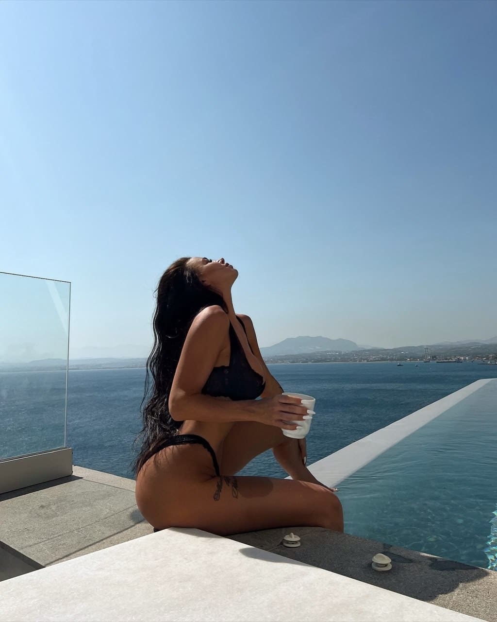 A woman in a black two-piece bikini sits on the edge of an infinity pool holding a white cup, head tilted back, with coastal scenery behind her.