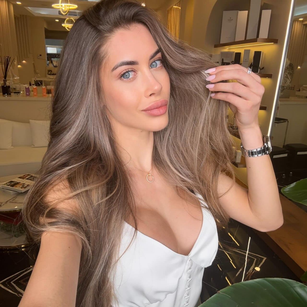 A woman in a white sleeveless top holds a strand of hair indoors, with shelves and a sofa visible in the background.