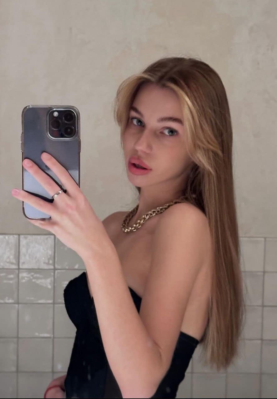 A blonde woman takes a mirror selfie in a black strapless top and gold chain against a tiled wall.