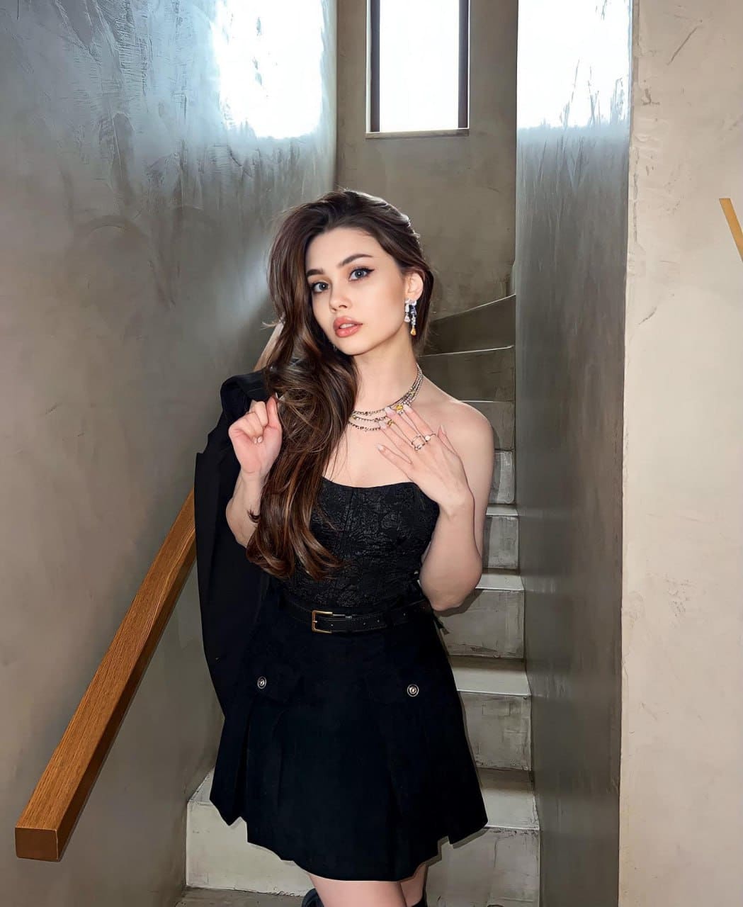 Mia stands on a concrete staircase in a black lace top, mini skirt, and jacket draped over one shoulder, wearing layered necklaces.