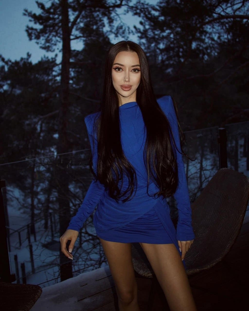 On an outdoor deck at dusk, she stands in a fitted blue mini dress with structured shoulders and bare legs.