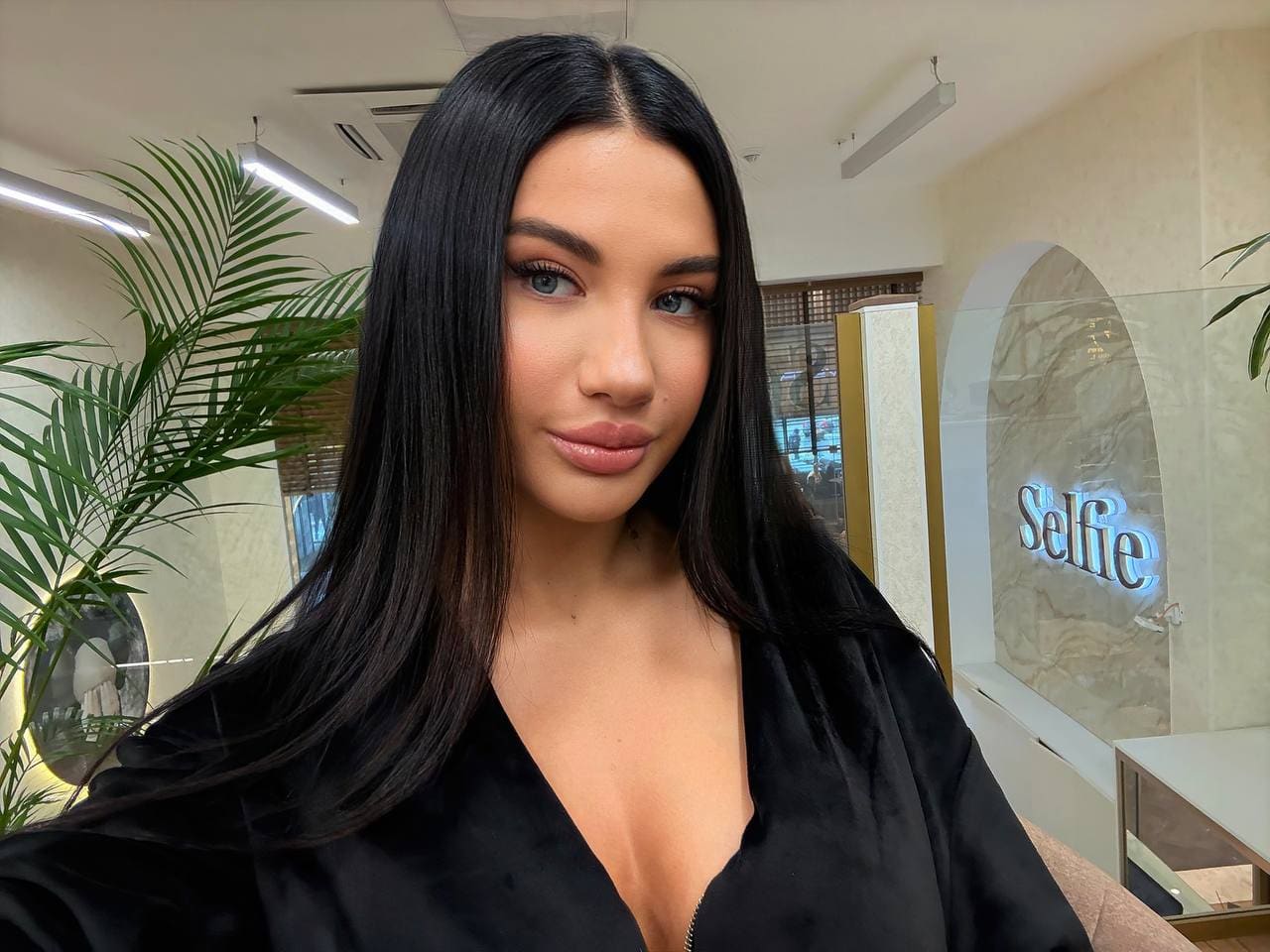 Margarita with long black hair and blue eyes wears a black deep-V top indoors near a potted plant and lit sign.