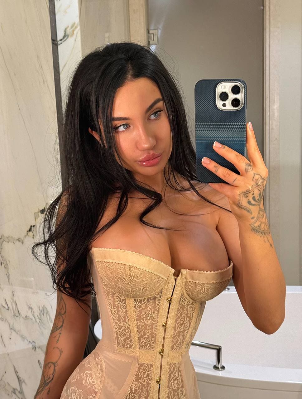 Mirror selfie in a bathroom showing a woman with arm tattoos wearing a beige lace corset with a deep neckline.