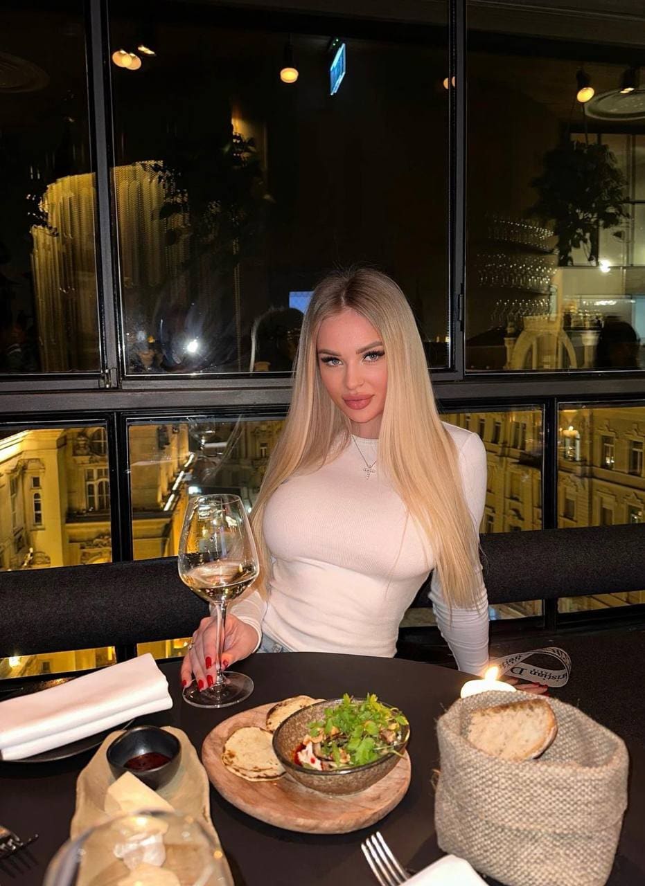 A woman in a fitted white long-sleeve top sits at a candlelit dinner table, holding a glass of white wine.