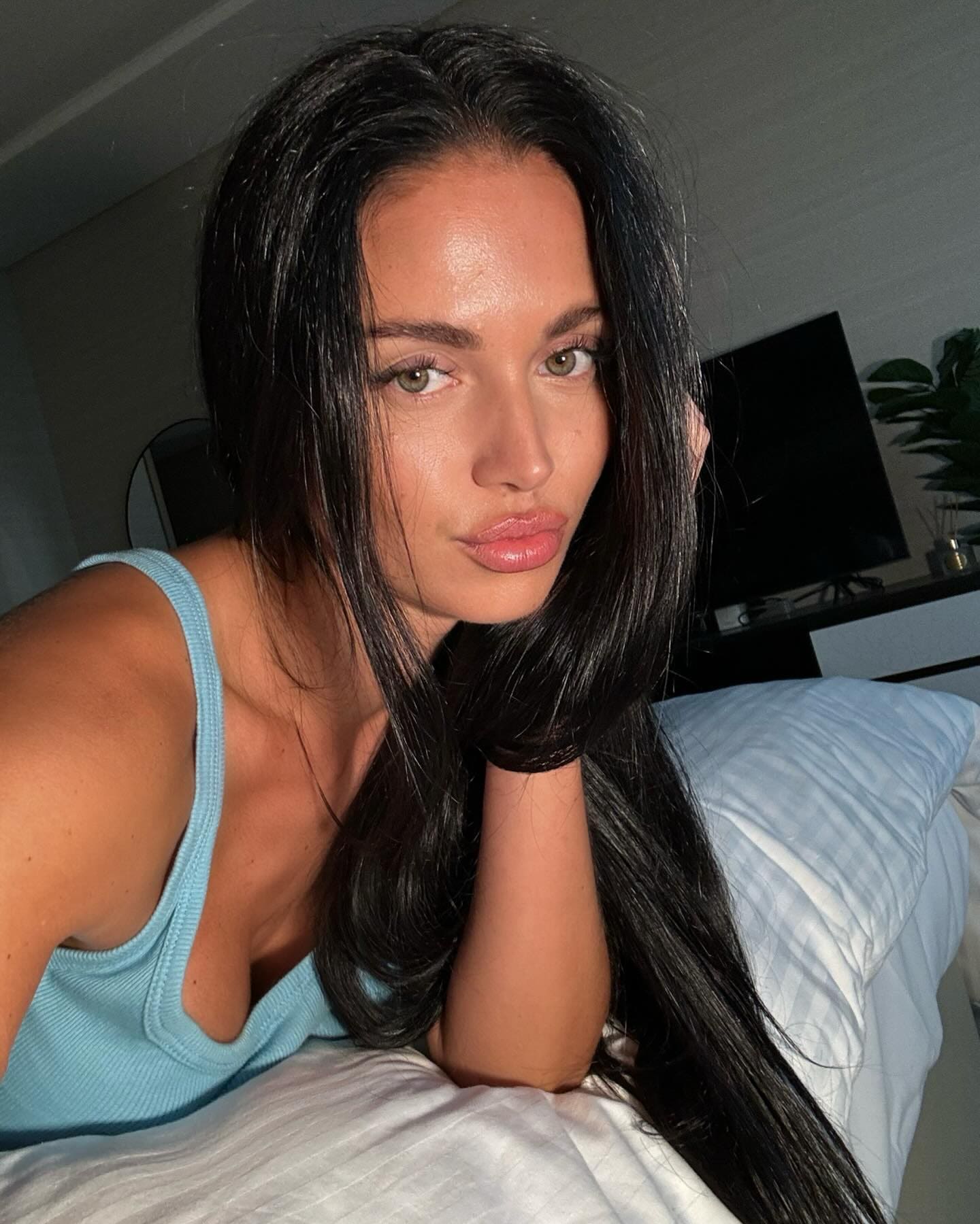 A woman lies on a bed with white linens wearing a light blue ribbed tank top, with a television and plant in the background.