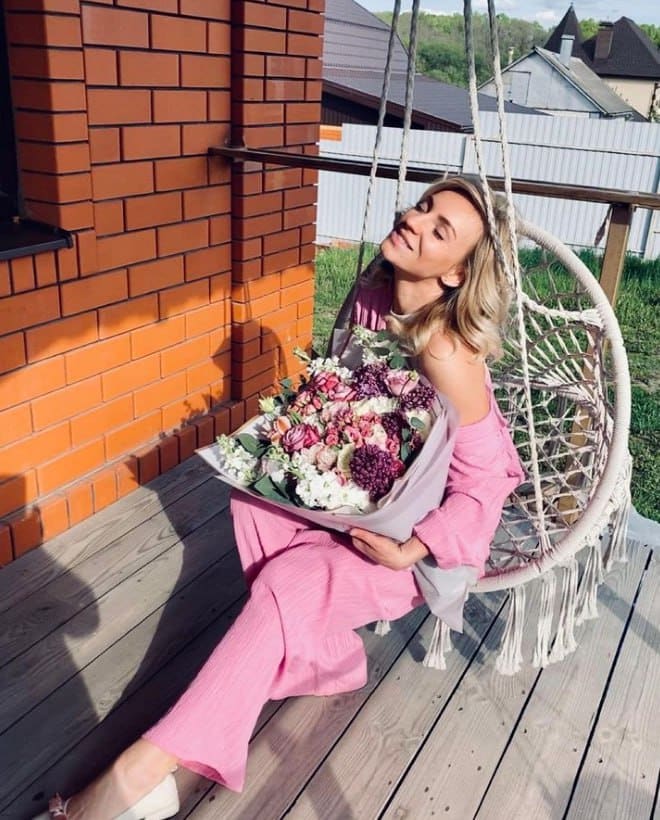 A blonde woman in a pink off-shoulder top and wide-leg trousers sits in a white macrame chair on a deck, holding a large bouquet.