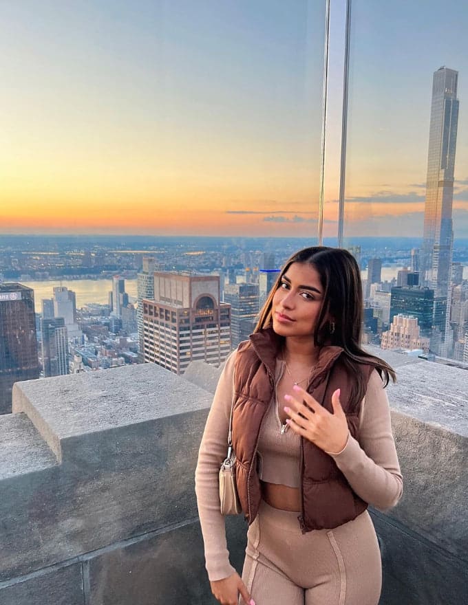 Standing on a rooftop terrace at sunset, a woman wears a brown puffer vest over a beige crop top and leggings, with a city skyline behind her.