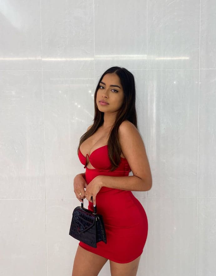 Victoria stands in a side pose against a white tiled wall, wearing a short red strapless dress with a chest cutout and holding a black handbag.