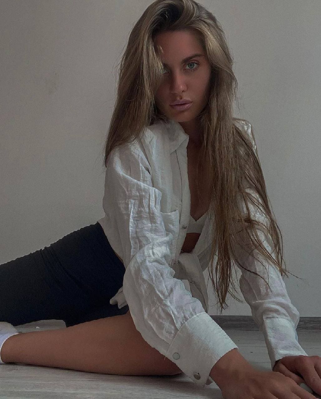 Rozaliya sits on a wooden floor against a gray wall, wearing an unbuttoned white shirt and black shorts with legs extended.