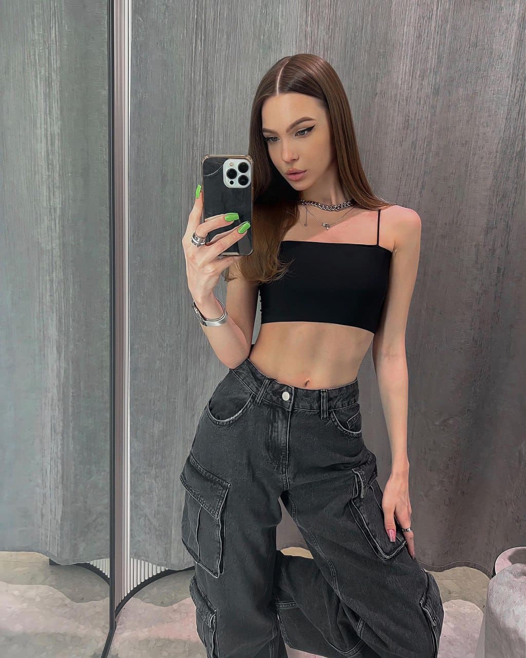 A mirror selfie in a gray room features a woman in a black crop top and cargo jeans, holding a phone with green nails, exposing her midriff.