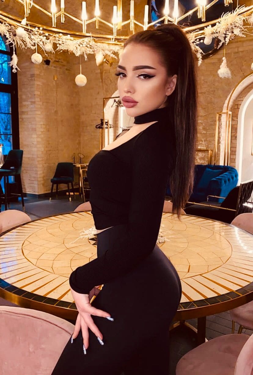 Profile view of a woman in a black cutout top beside a mosaic table, with brick walls and hanging lights in the background.