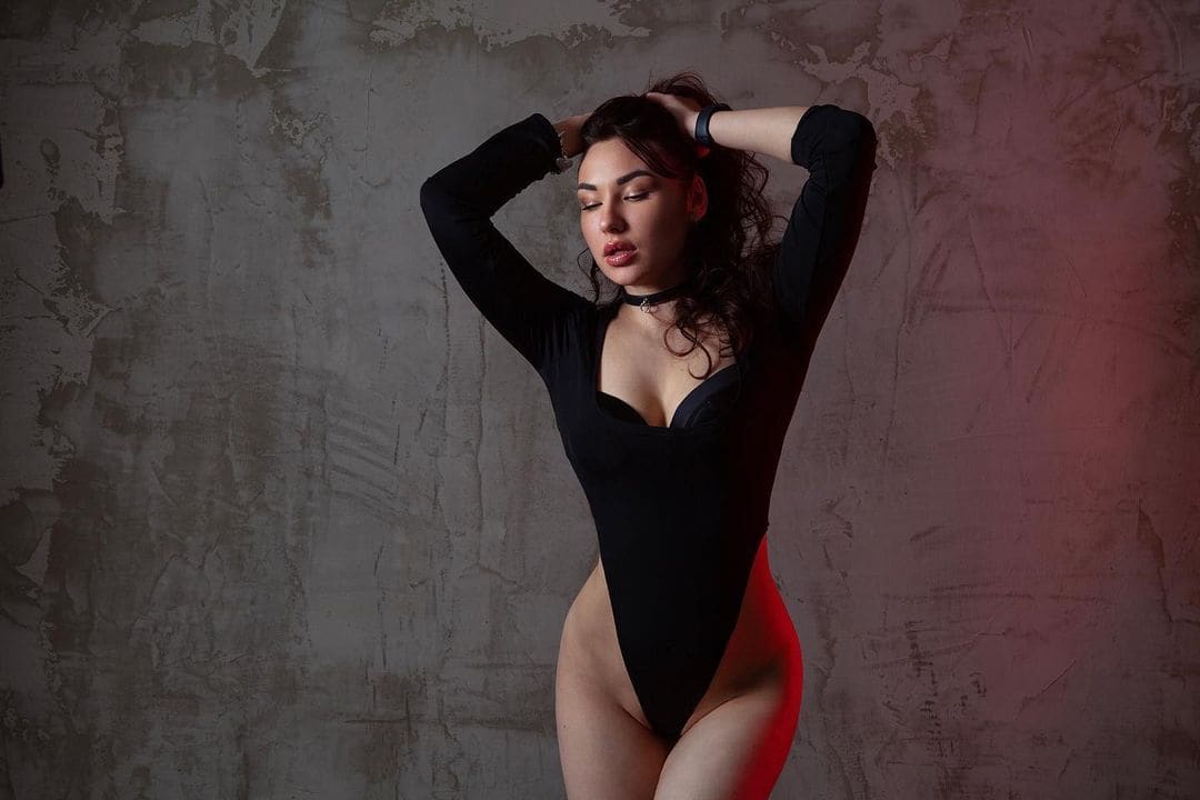 A woman stands with arms behind her head, wearing a black high-cut bodysuit and choker, against a gray wall with red lighting.