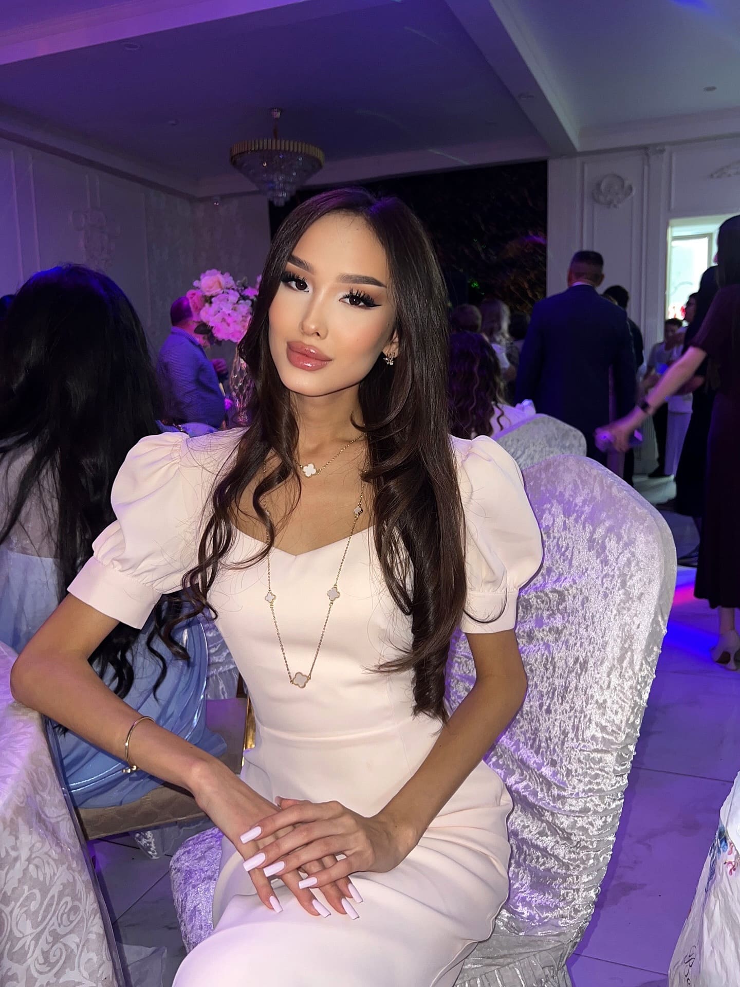 Gulzira sits in a silver chair at an event, wearing a light V-neck dress and pendant necklace, with guests standing under purple lighting.