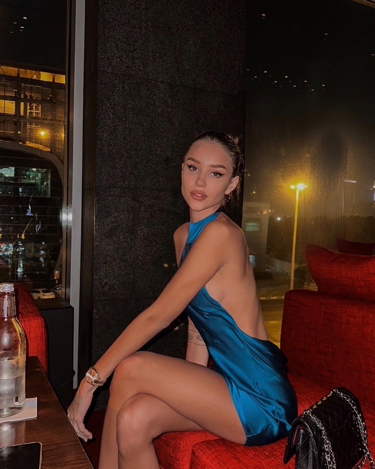 A woman in a teal satin dress sits on a red bench in a dim room, legs crossed, next to a glass bottle.
