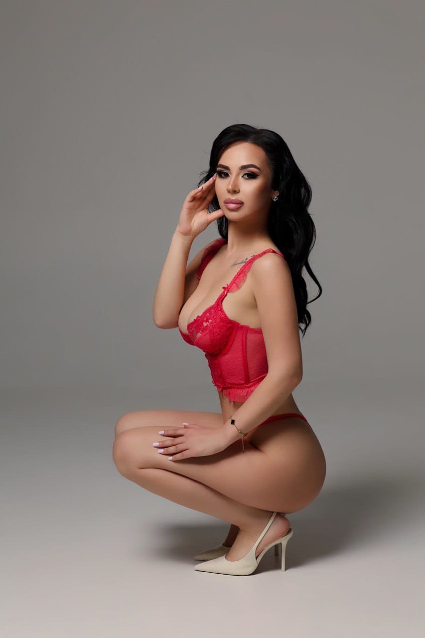 A woman crouches in a gray studio wearing a red lace bodysuit and white heels, her hand raised to her temple.
