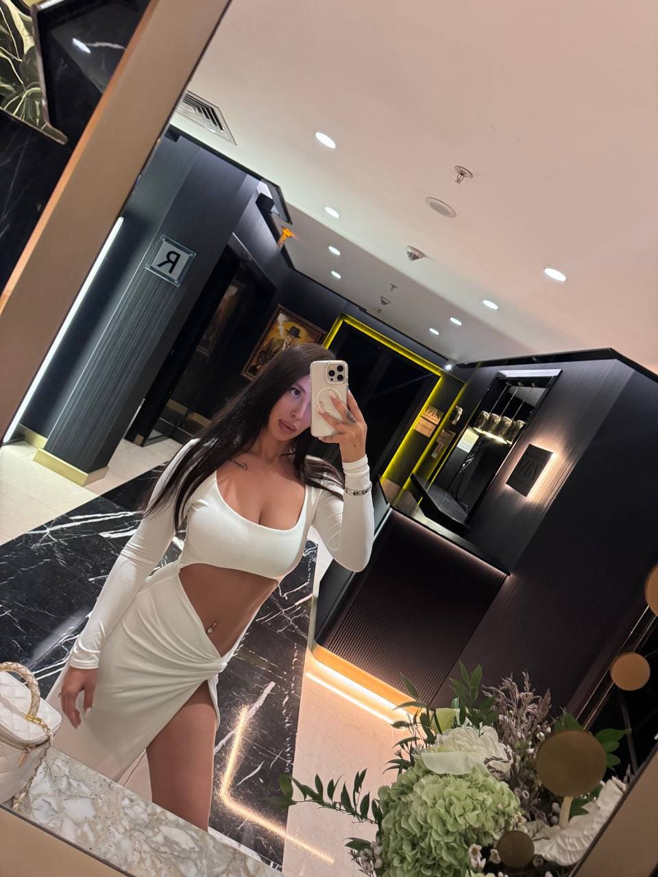 A woman stands in a modern interior wearing a white crop top and high-slit skirt, with marble floors and flowers nearby.