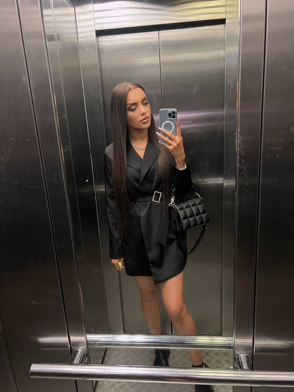 Susie in a black belted blazer dress and boots, taking a mirror selfie in a stainless-steel elevator with a quilted bag.