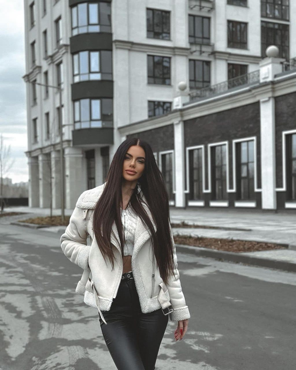 A woman stands on a street before a modern building, wearing a white shearling-lined jacket, cropped top, and black pants.
